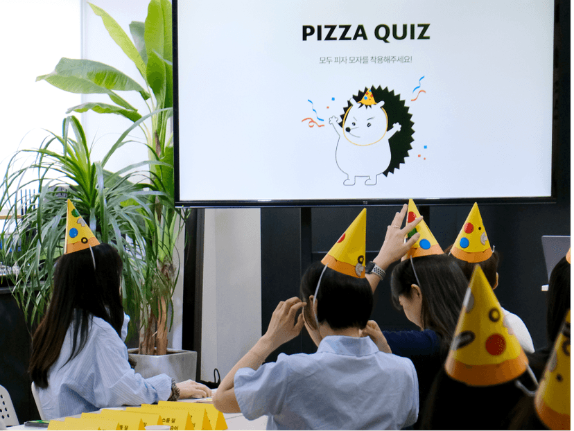 사람들이 피자 모양의 종이 모자를 쓰고 퀴즈를 풀고 있다. 중앙의 스크린에는 "PIZZA QUIZ"라는 글자와 고슴도치 캐릭터가 있다.